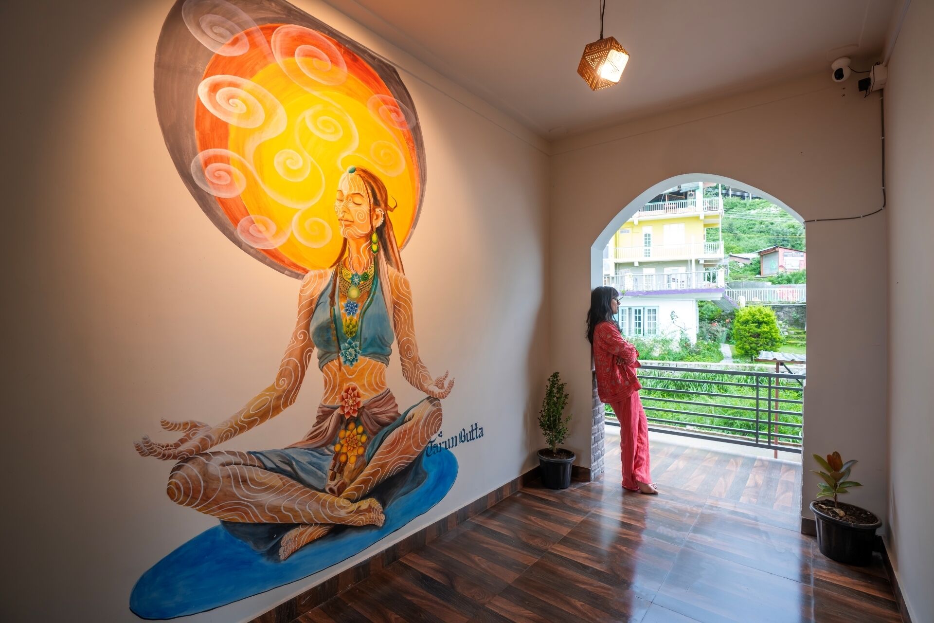 Una habitación con un mural de una figura meditativa, un balcón con vistas a edificios y una planta en maceta.