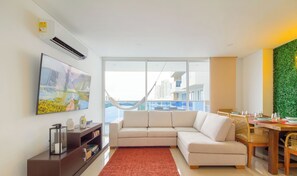Ein modernes Wohnzimmer mit einer Couch, einem Esstisch mit Stühlen, einem großen Fenster mit Blick auf Gebäude und einer Hängematte.