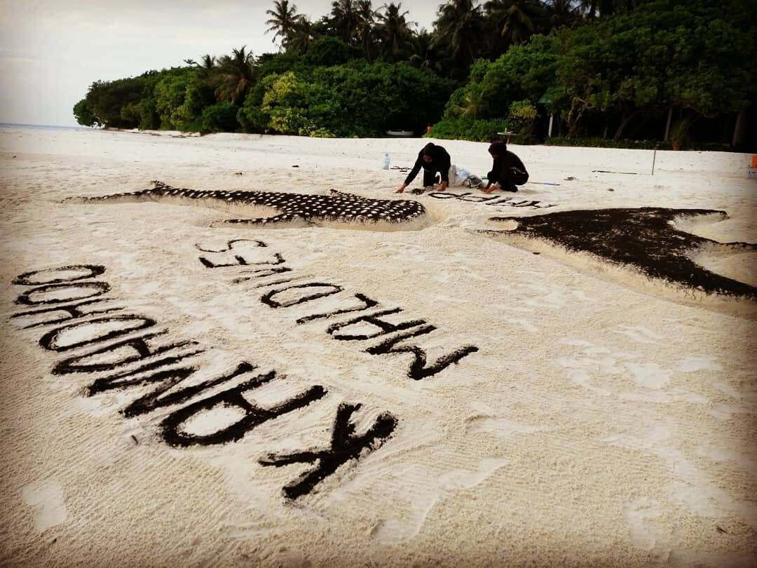 A beach with sand art spelling out 'KIRINDU' and two people in the background.