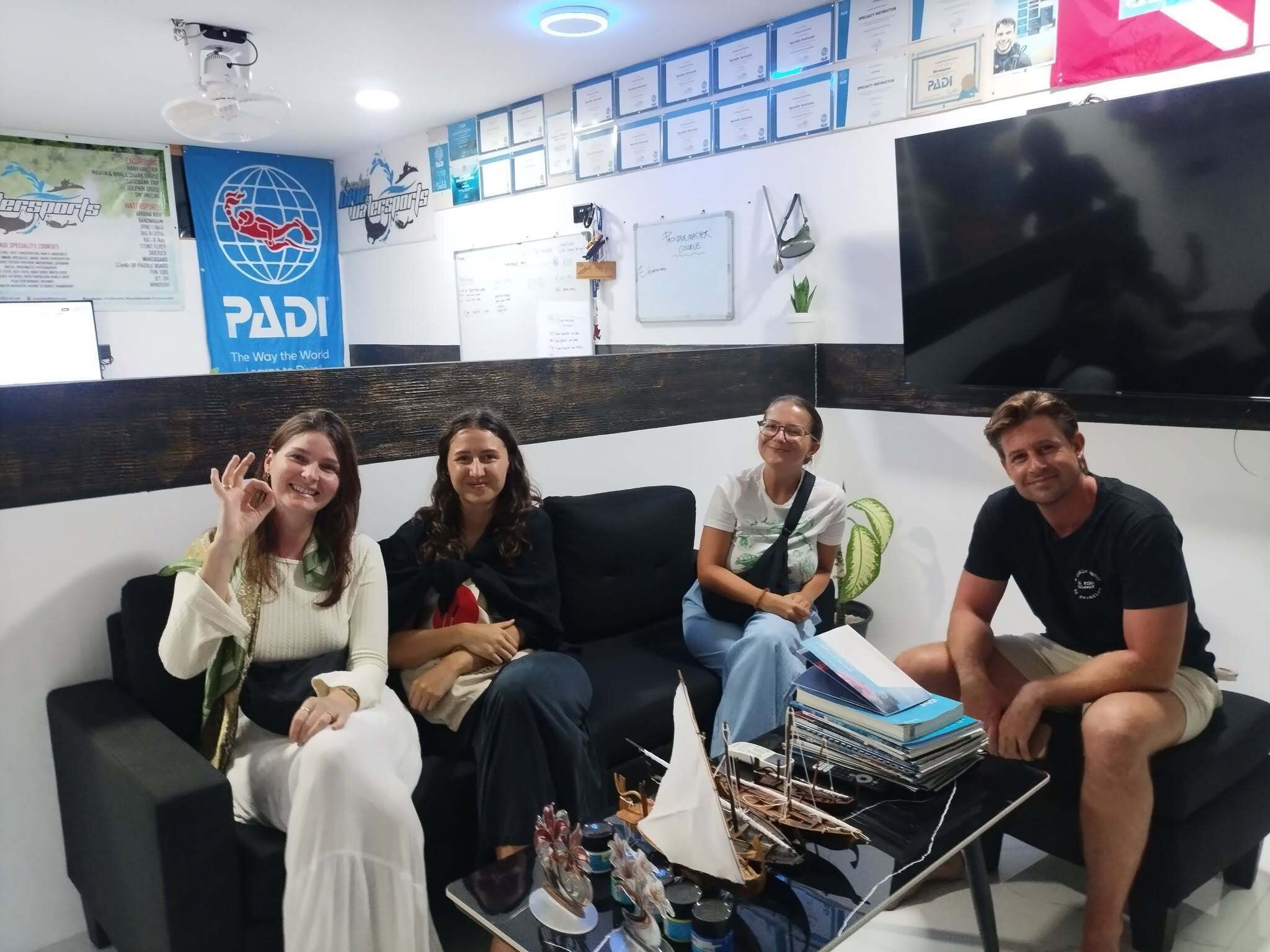 Un espace d’accueil avec un canapé noir, une table basse sur laquelle sont posés des livres et une maquette de bateau, et un mur orné de certificats encadrés et d’un grand logo PADI.