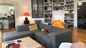 Una sala de estar con un sofá gris, una mesa de centro con libros y una tableta, una estantería llena de libros y fotografías enmarcadas, y una lámpara roja sobre una mesita auxiliar.