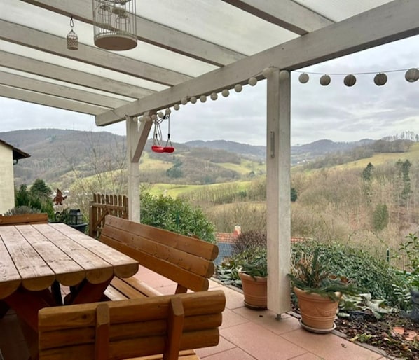 A covered patio with a wooden table and benches, potted plants, and a view of rolling hills.
