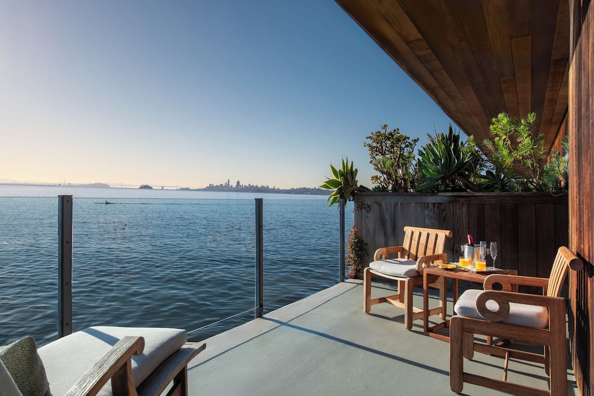 Un balcón con vista al agua, sillas de madera, una mesa con bebidas y una pared cubierta de plantas.