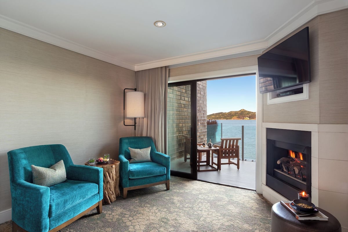 Habitación de hotel con chimenea, dos sillones azul verdosos, una pequeña mesa con una vela y vista al mar a través de un ventanal de vidrio.