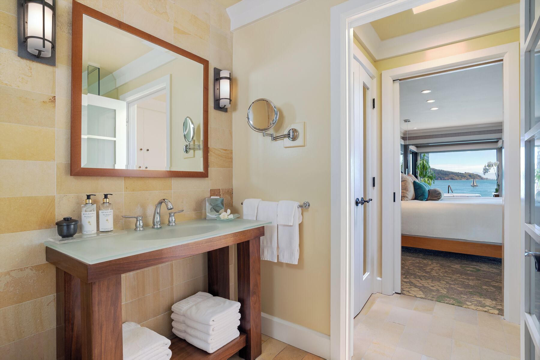 Un baño con tocador de madera, un espejo grande, dos luces fijadas a la pared y vistas a un dormitorio con cama y ventana con vista al agua.