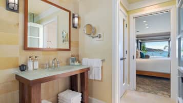 Un baño con tocador de madera, un espejo grande, dos luces fijadas a la pared y vistas a un dormitorio con cama y ventana con vista al agua.
