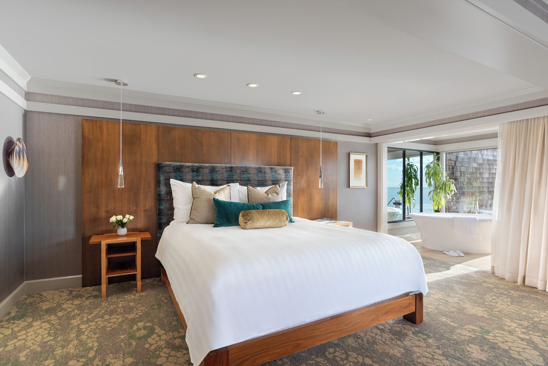 A hotel room with a large bed, a wooden headboard, a nightstand, a potted plant, and a view of the sea through a large window.