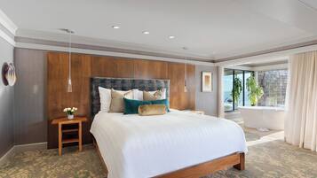 A hotel room with a large bed, a wooden headboard, a nightstand, a potted plant, and a view of the sea through a large window.