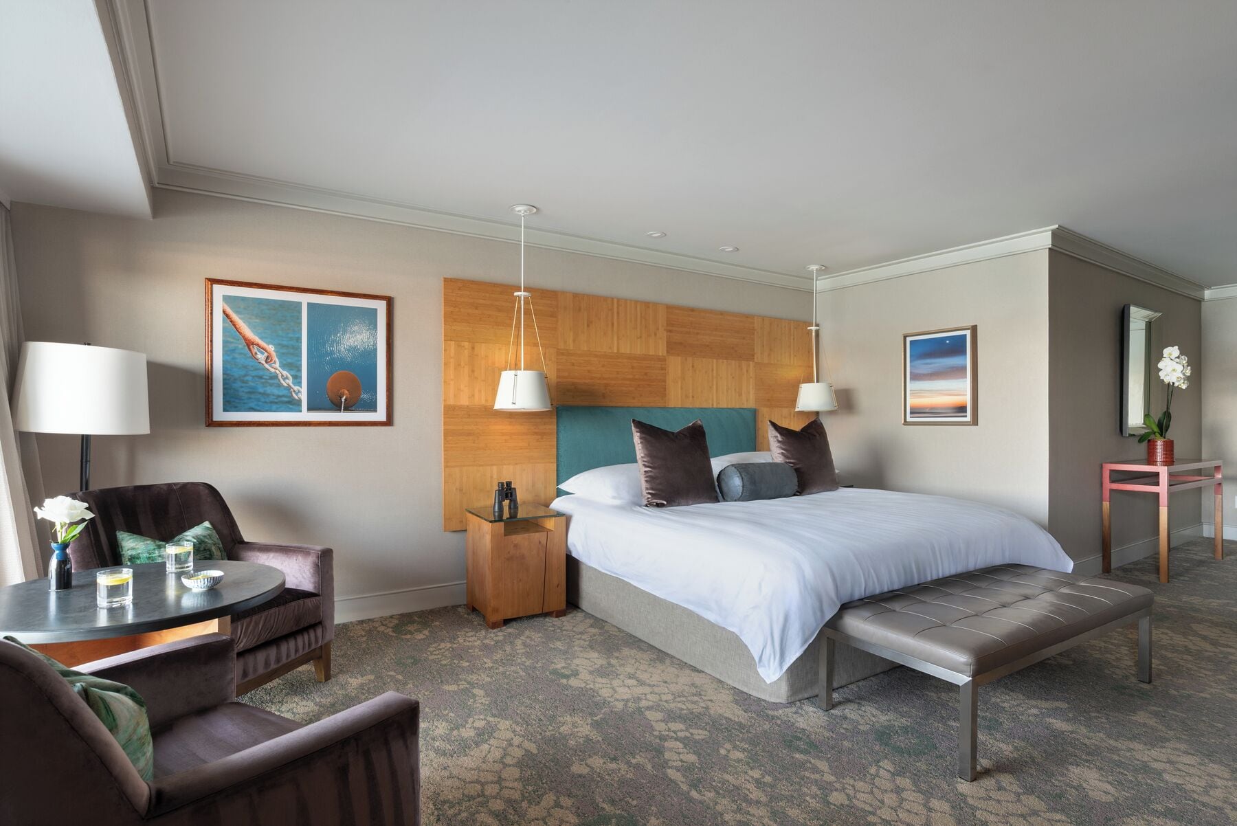 A hotel room with a bed, bedside tables, a desk, a chair, a bench, and artwork on the walls.