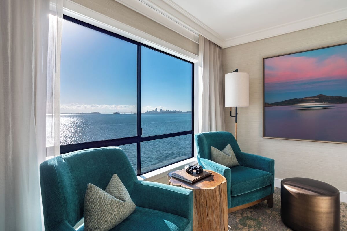Una habitación con dos sillones turquesa, una mesita redonda de madera, una lámpara de pie, un cuadro y un amplio ventanal con vista al horizonte de la ciudad.