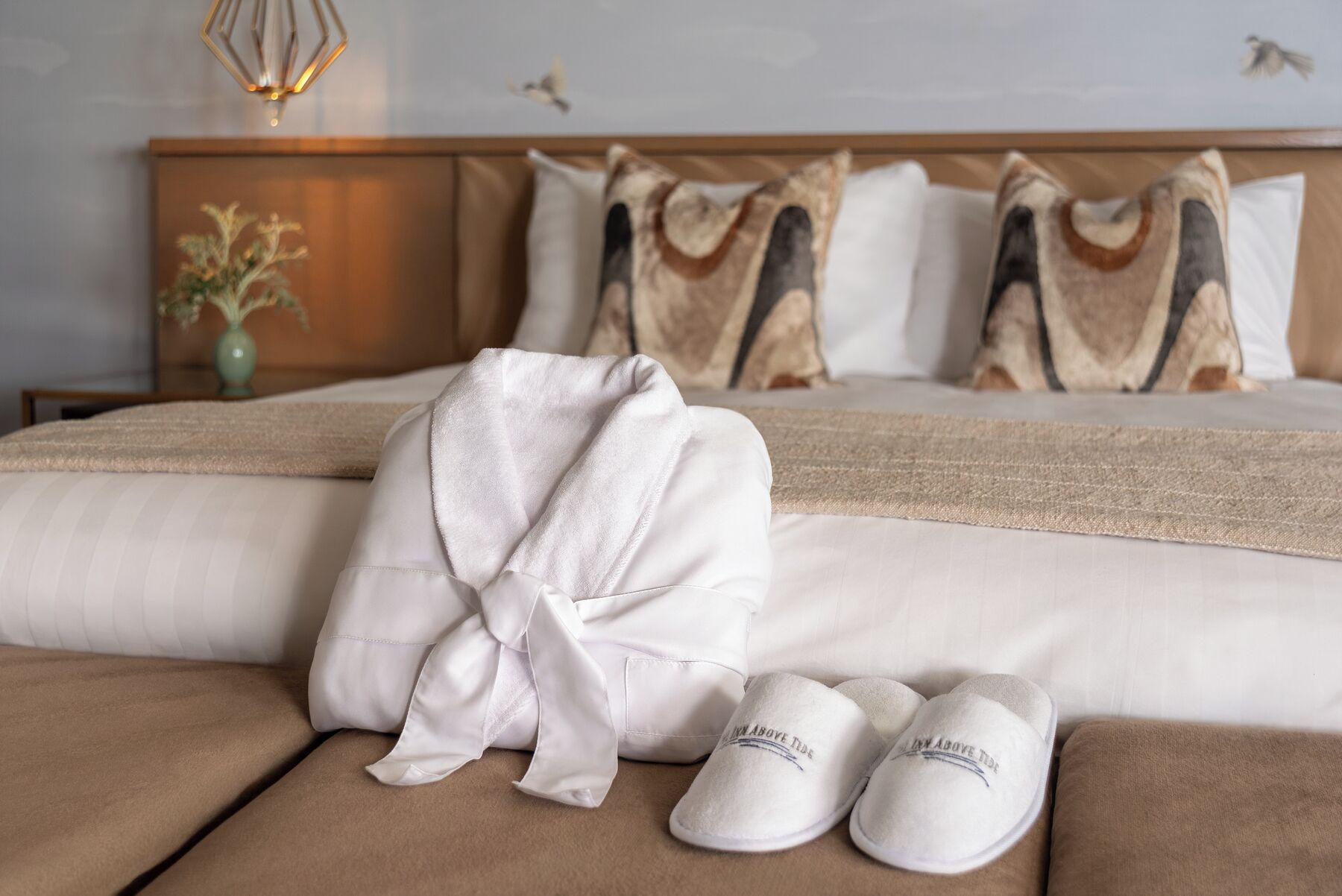 A neatly made bed with a white robe and slippers, a vase with flowers, and a bird on the wall.