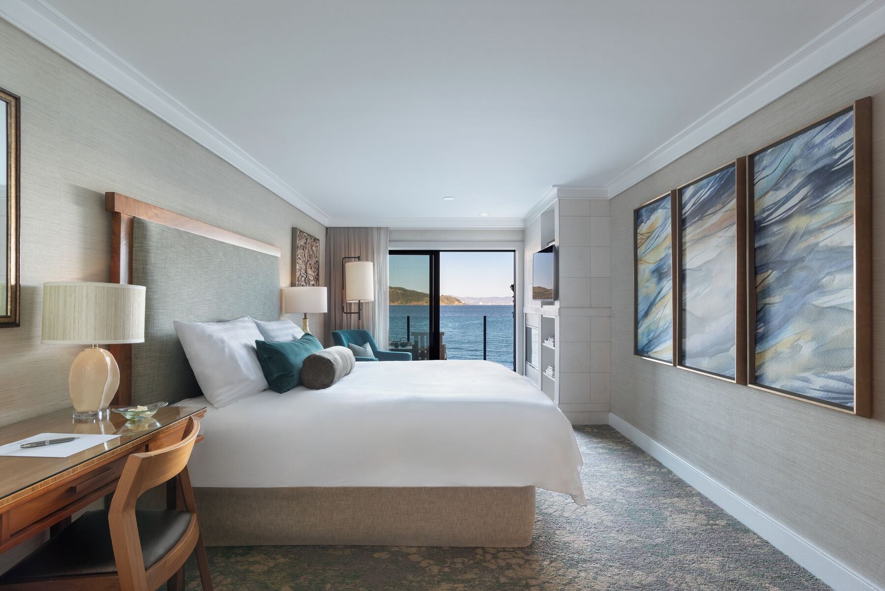 A hotel room with a large bed, a desk, a chair, a TV, and a view of the sea.