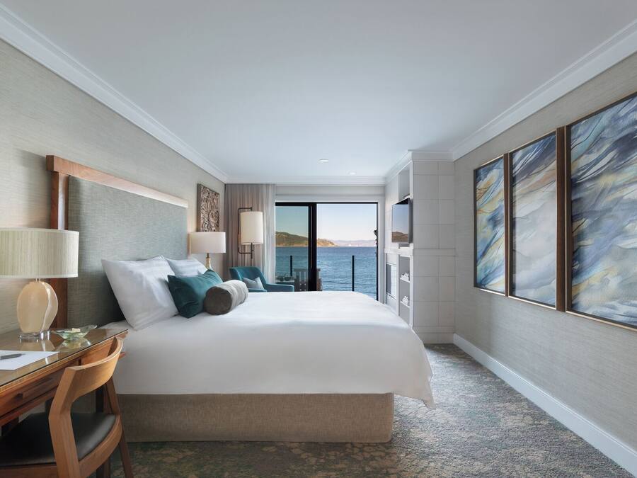 A hotel room with a large bed, a desk, a chair, a TV, and a view of the sea.