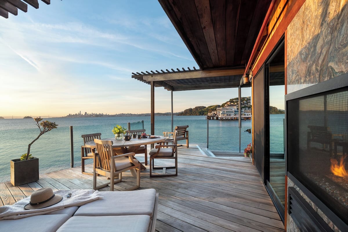 Una terraza de madera con una mesa, sillas, un sofá, una chimenea y vistas al agua y al horizonte de la ciudad.