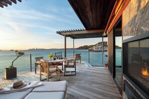 Una terraza de madera con una mesa, sillas, un sofá, una chimenea y vistas al agua y al horizonte de la ciudad.