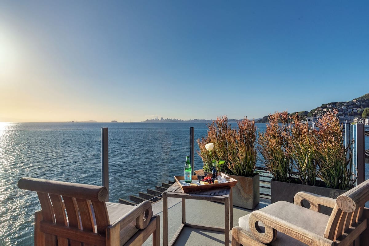 Una terraza con sillas de madera, una mesa con una bandeja de comida y bebidas, y vistas al mar y al horizonte de la ciudad.