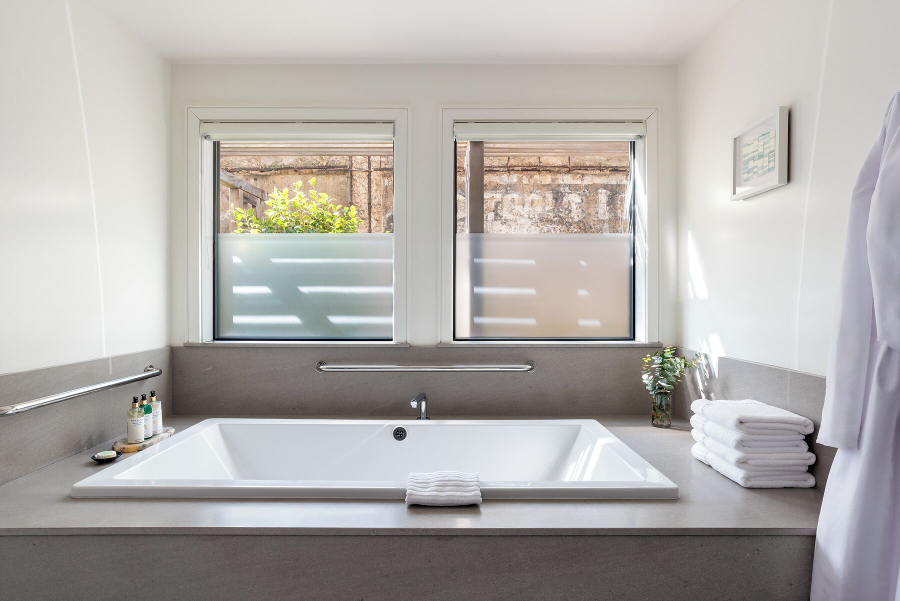 A modern bathroom with a large bathtub, two windows, a towel rack, and a small plant.