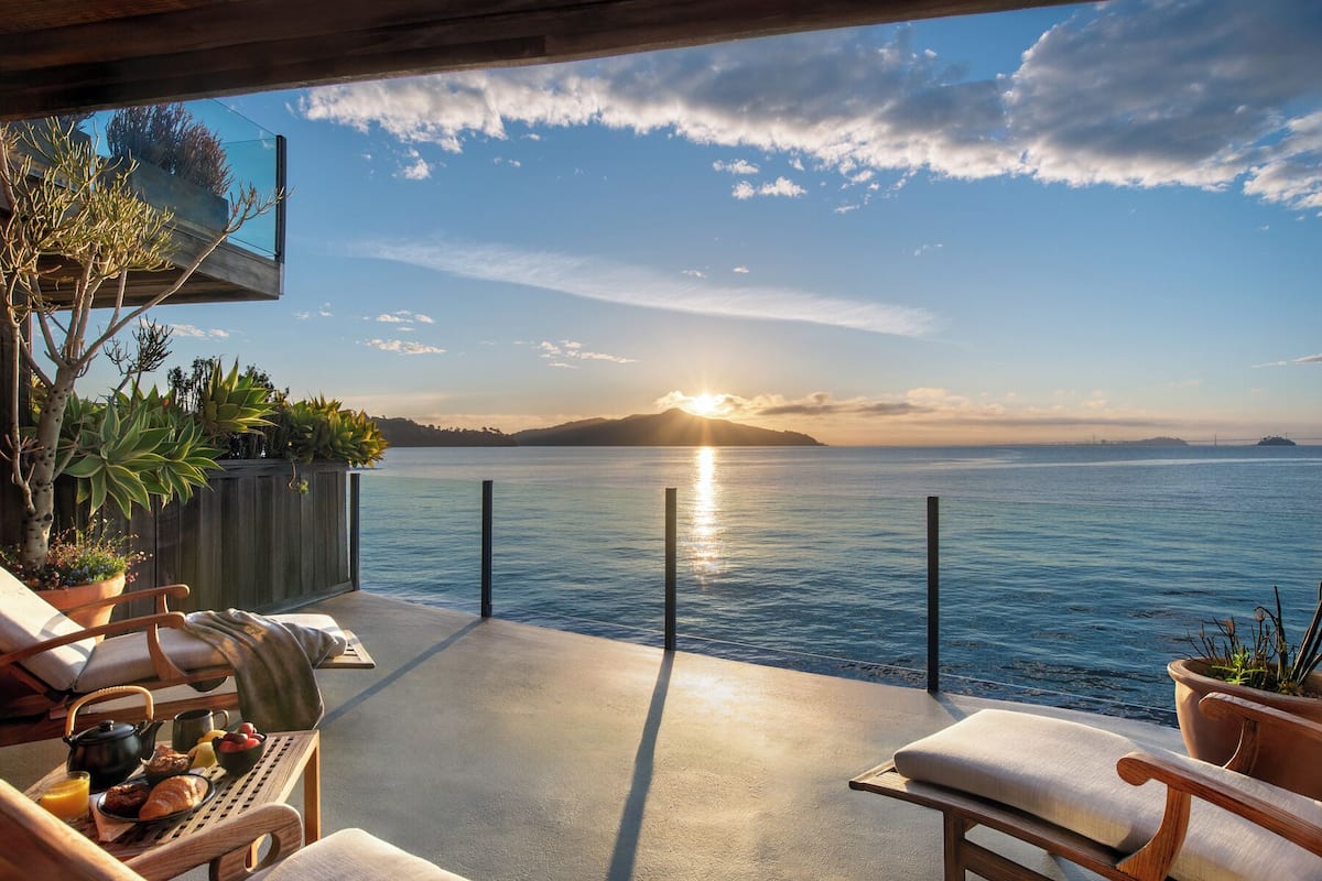 Un balcón con vista al mar, una mesa de madera con desayuno y plantas en macetas.