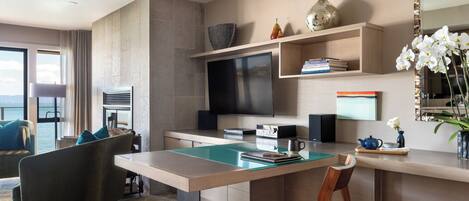 A modern hotel room with a dining area, a flat-screen TV, a bookshelf, and a view of the sea.
