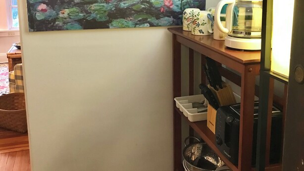 Et værelse med et maleri af en sø, kaffemaskine og køkkenudstyr på hylder.