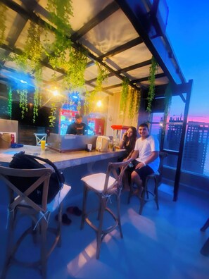 Un bar en la azotea con plantas colgantes, un refrigerador de Coca-Cola y vistas de la ciudad al atardecer.