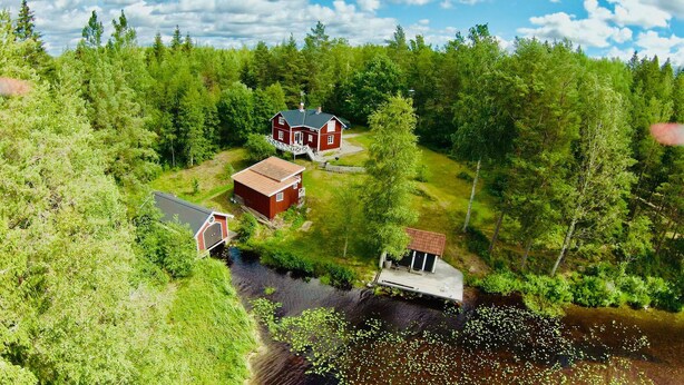Una casa de madera roja con porche, rodeada de un bosque denso y un pequeño cuerpo de agua.