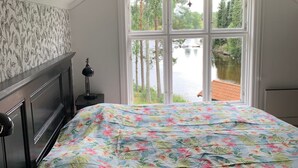 Un dormitorio con cama, mesita de noche, lámpara y ventana con vistas a árboles y un cuerpo de agua.
