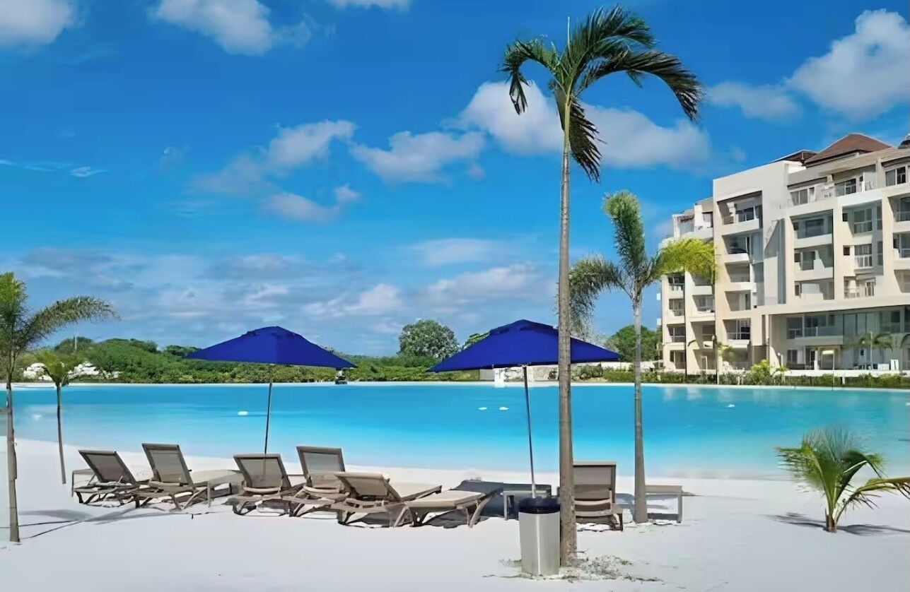 A resort pool area with lounge chairs and umbrellas, surrounded by palm trees and a building with multiple balconies.