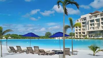 A resort pool area with lounge chairs and umbrellas, surrounded by palm trees and a building with multiple balconies.