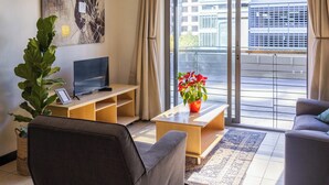 Ett vardagsrum med en soffa, ett soffbord, en tv, en stor växt och en skjutdörr i glas som leder ut till en balkong.