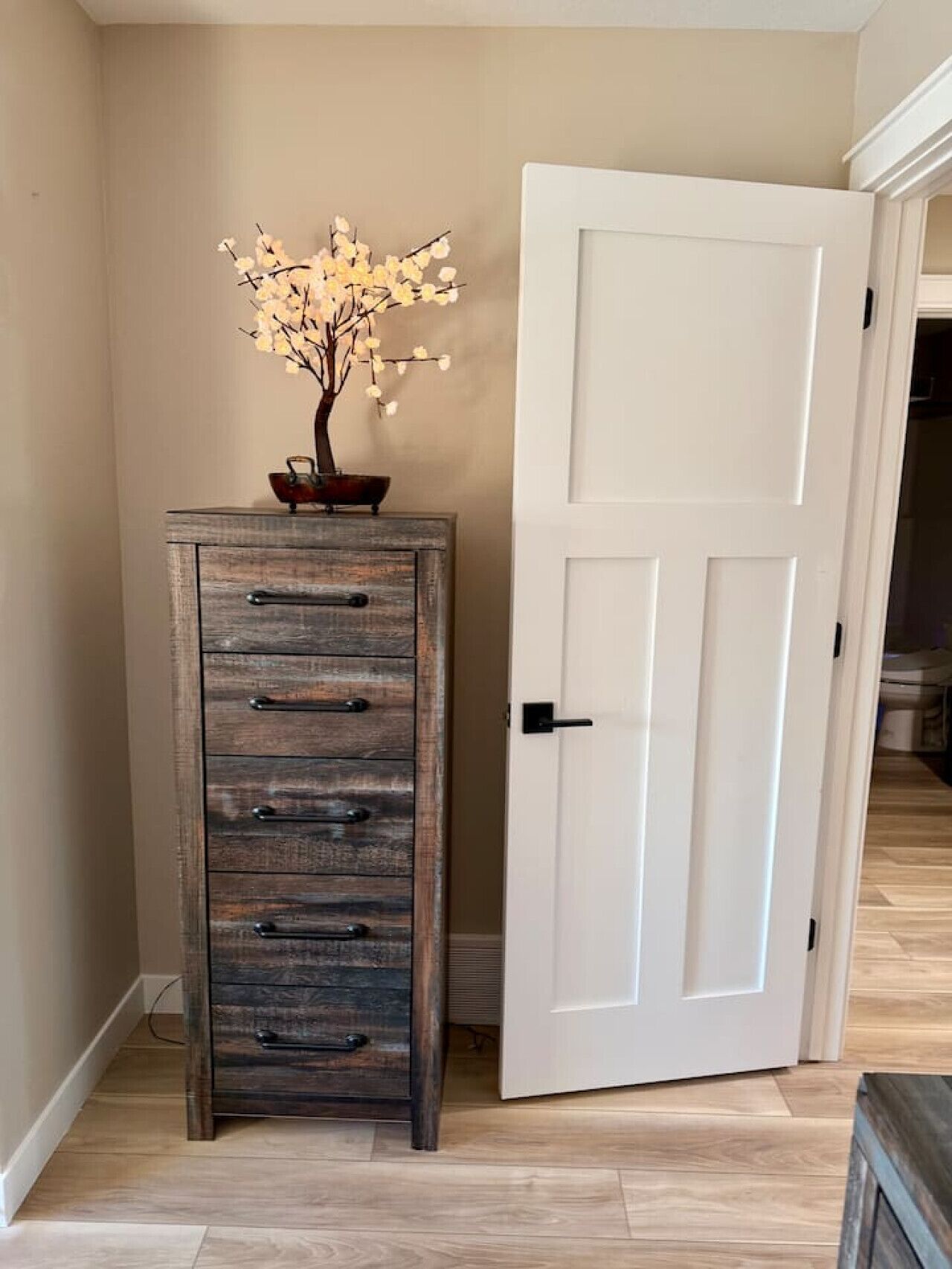 Une commode en bois avec des tiroirs et un vase de fleurs, une porte blanche avec une poignée noire, et un plancher en bois.