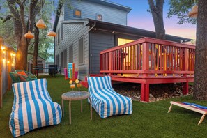 Un patio trasero con una terraza, dos sillones colgantes a rayas, una mesita con bebidas y una casa al fondo.
