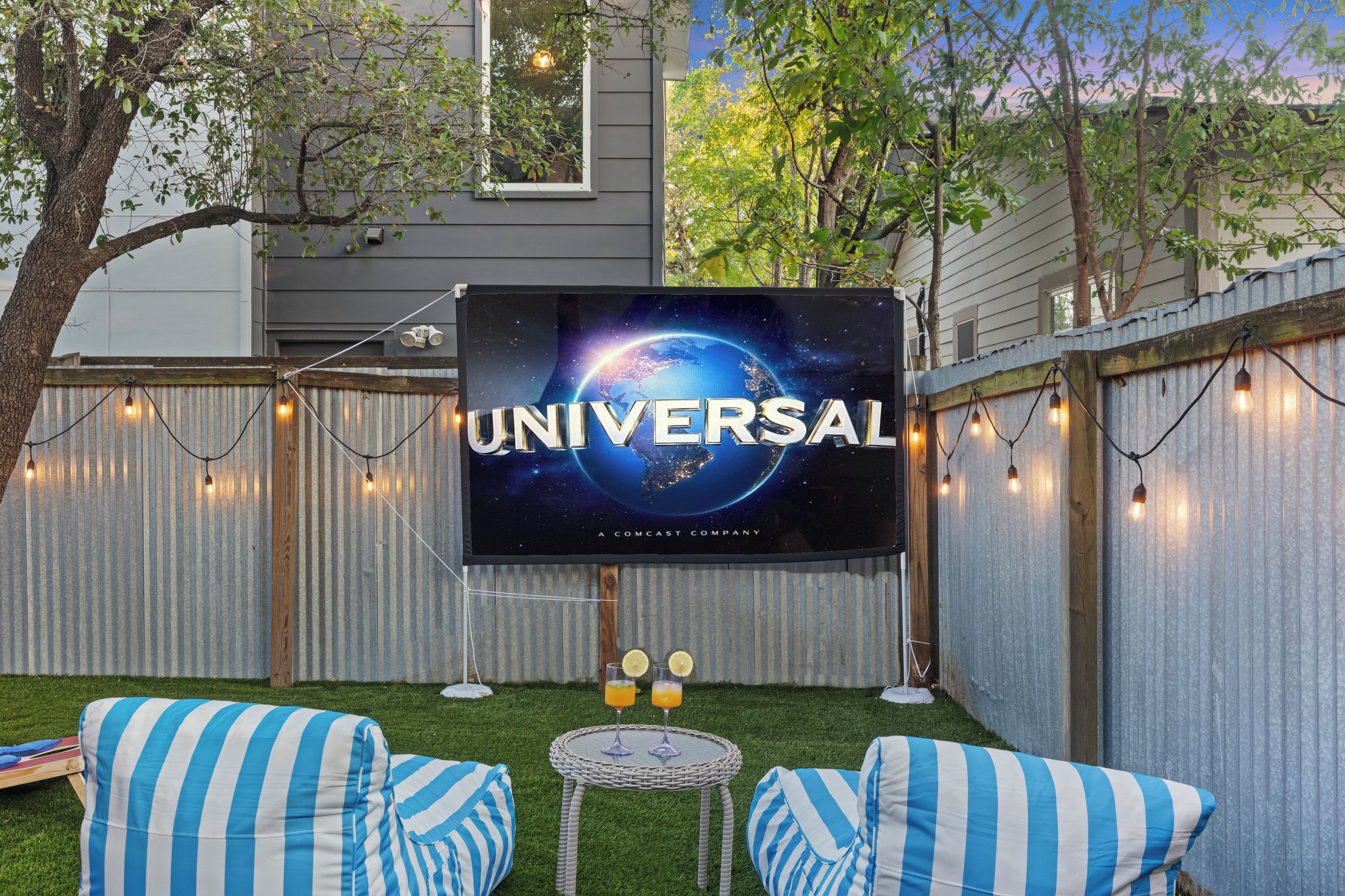 A backyard with a large screen displaying the Universal logo, a small table with drinks, and striped cushions.