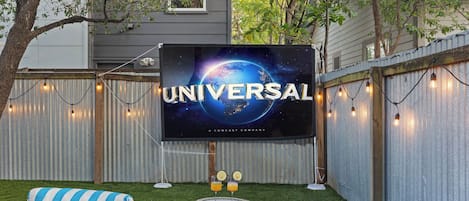 A backyard with a large screen displaying the Universal logo, a small table with drinks, and striped cushions.