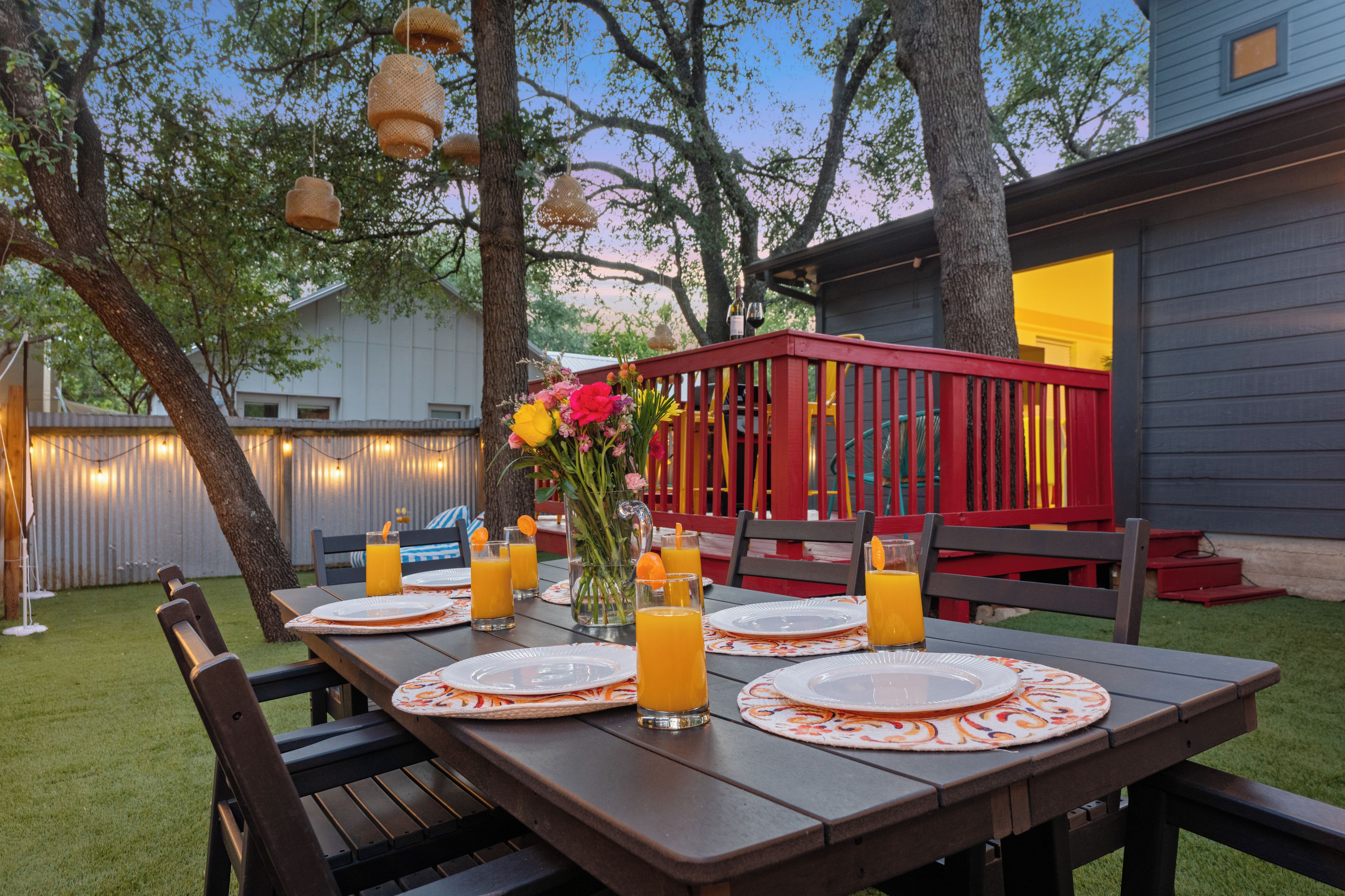 An outdoor dining area with a wooden table set for four, surrounded by chairs, and a red deck in the background.