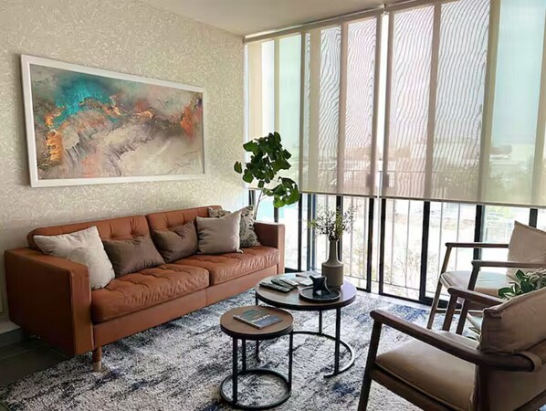A living room with a brown sofa, a round coffee table, a potted plant, and a large window with blinds.