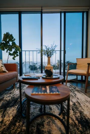 A modern living room with a sofa, two coffee tables, a potted plant, and a view through large windows.