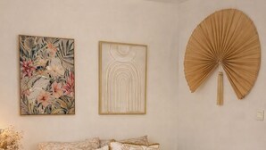 A bedroom with a bed, two wall-mounted artworks, a wall fan, and a bedside table with a vase. - Cosy furnished studio, quiet, nature (Saint-Joseph)