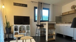 A modern living room with a flat-screen TV, a dining area with a table and chairs, a kitchen with white cabinets, and a large window with curtains. - Cosy furnished studio, quiet, nature (Saint-Joseph)