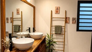 A modern bathroom with two sinks, a large mirror, wooden vanity, a plant, and a window with blinds. - Cosy furnished studio, quiet, nature (Saint-Joseph)