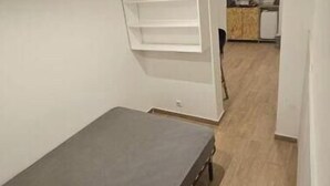 Ett rum med en säng, en vit hylla och ett litet kök i bakgrunden.