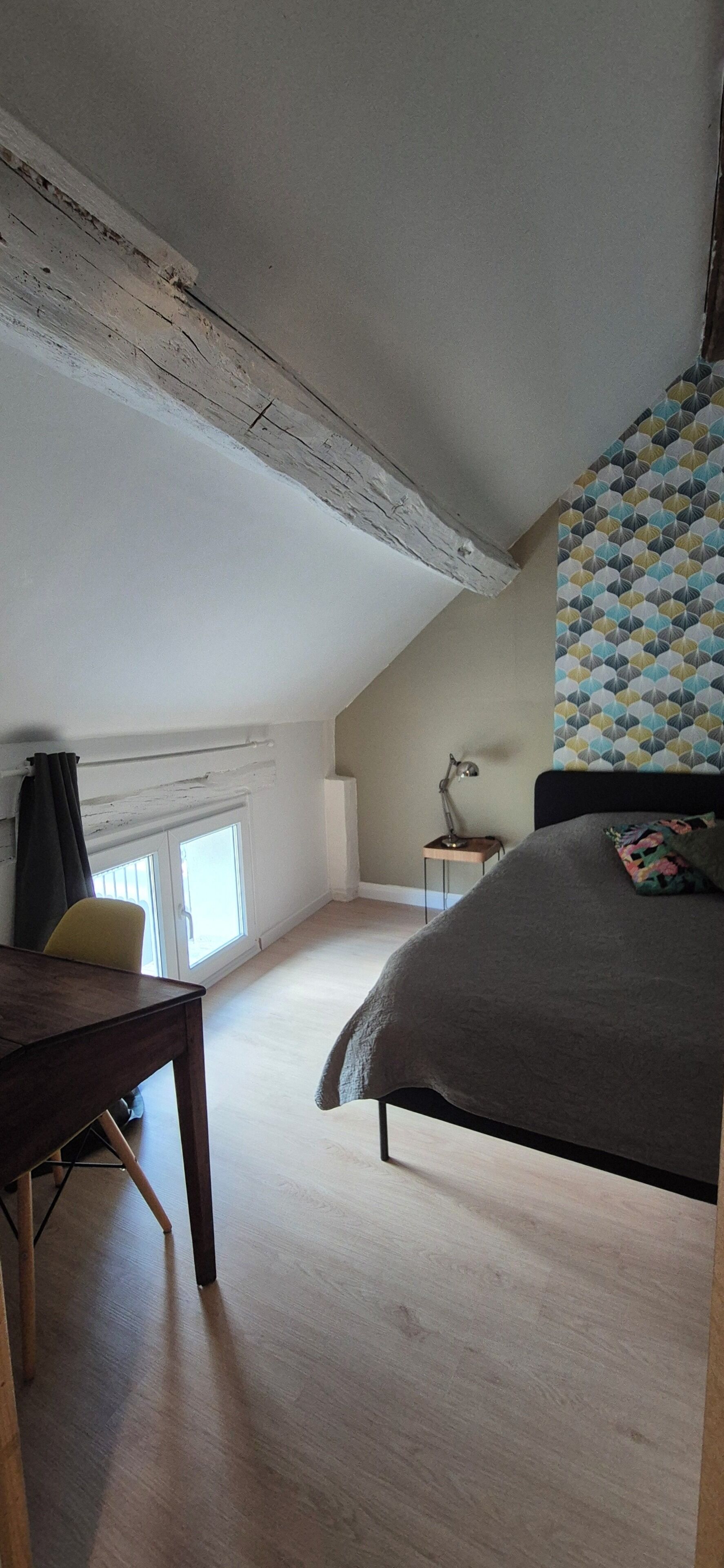 A room with a sloped ceiling, a bed with a patterned headboard, a desk with a chair, a small table, and a lamp.