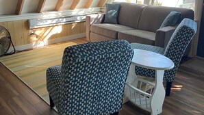 A cozy attic room with a wooden sloped ceiling, a sofa, patterned chairs, a small table, and a fan. - Cozy 2-bedroom cottage in charming Pierson, Michigan! (Pierson)