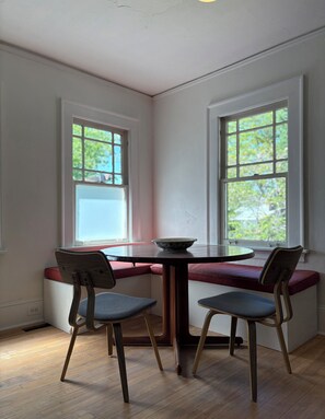 Un rincón comedor con una mesa redonda y dos sillas, un asiento junto a la ventana, y vista a zonas verdes al exterior.