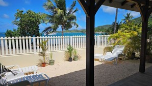 Un espace extérieur aménagé, abrité et doté de chaises et de tables en plastique blanc, entouré de plantes en pot et d’une clôture blanche, offrant une vue sur une plage et l’océan.