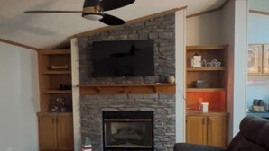 A living room with a stone fireplace, wooden flooring, a ceiling fan, and built-in shelving. - Lakefront Home-Beautiful Views & Private Dock 15-20 to Clemson, Anderson,Seneca (Townville)