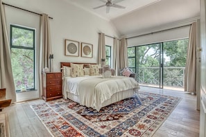 Une chambre spacieuse avec un grand lit, une commode en bois, un ventilateur de plafond et une vue sur la verdure grâce à de grandes fenêtres.