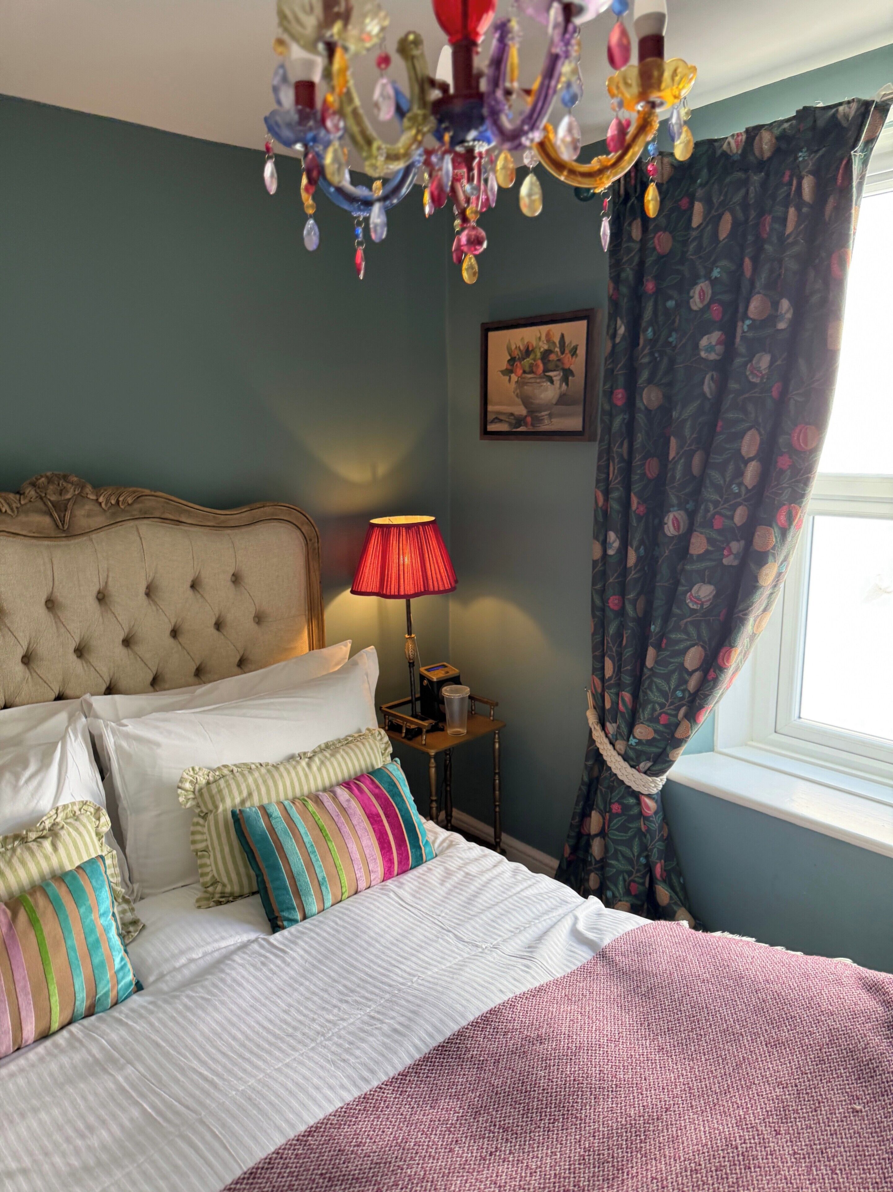 A bedroom with a tufted headboard, a bedside lamp, a framed picture on the wall, and a window with curtains.