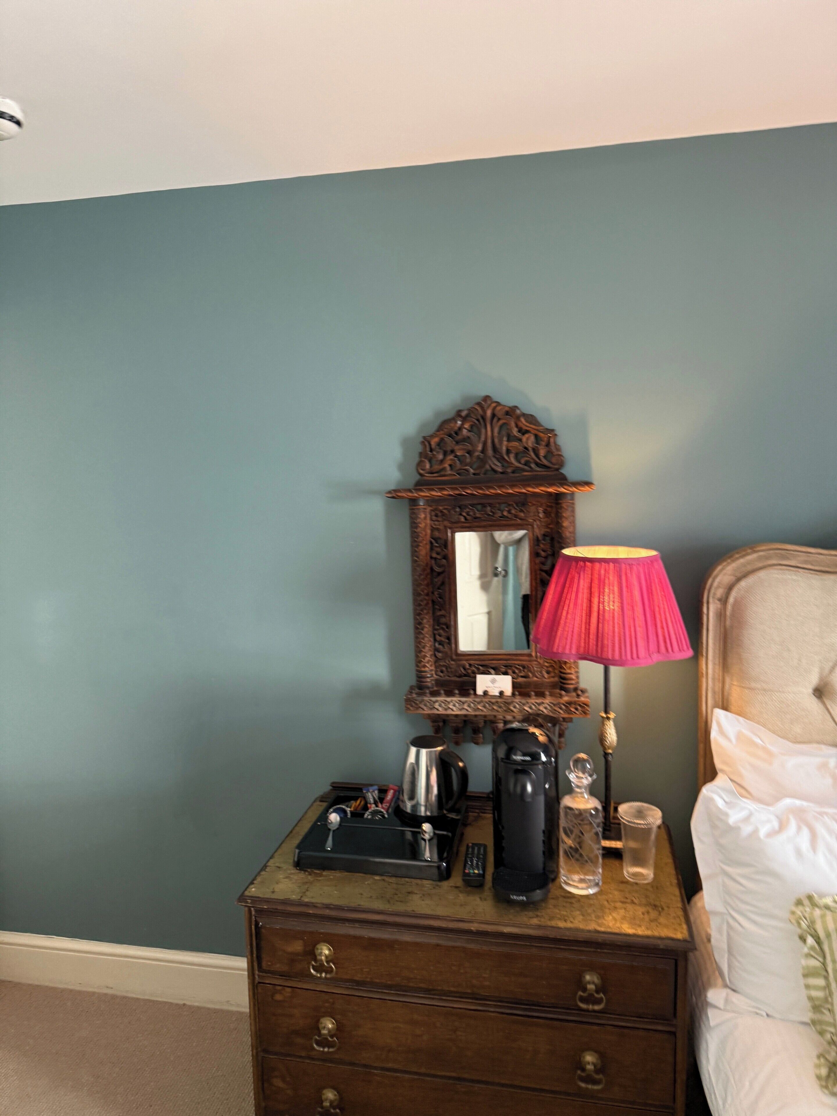 A bedroom with a bed, a dresser with a mirror, a lamp, and a nightstand with a coffee maker and remote controls.