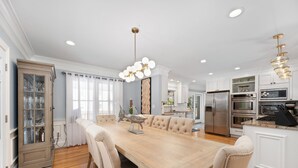 Un comedor con una mesa de madera, sillas tapizadas en color beige, una araña y una cocina con electrodomésticos de acero inoxidable.